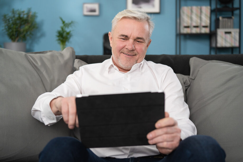 Smiling middle aged old man relaxing holding digital tablet reading e book sit on sofa at home, senior adult man enjoying using computer pad apps browsing internet shopping on couch in living room